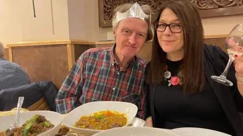 Tim Hartley and his wife Helen Lucitt are sitting at a table full of different curry dishes. Tim is sat on the left of Helen wearing a chequered blue and red shirt. He is wearing a silver paper crown from the crackers. Helen is lifting up a glass of white wine. She is wearing glasses and a black top.