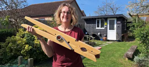 Steve Wainwright Cat Thompson has light brown hair and wears glasses, she is wearing a burgundy T-shirt and is holding a giant clothing peg which is about two feet (0.61m) long. She is in a sunny back garden, with a black shiplapped garden shed behind her on a grass lawn.