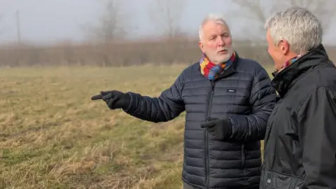 David Still and Nick Hayward, both in their 60s, are wearing dark jackets and colourful snoods. They're standing talking and gesturing in a field and there is a misty grass verge behind them.