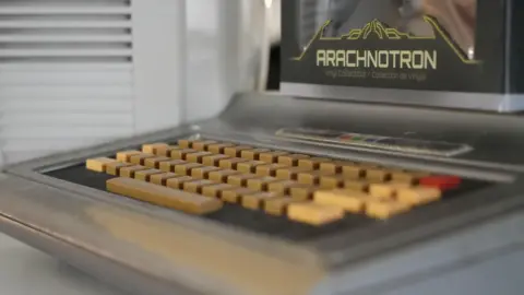 Shaun Whitmore/BBC A close up of a retro game. There is writing on the game which says, ARACHNOTRON on it in yellow writing.