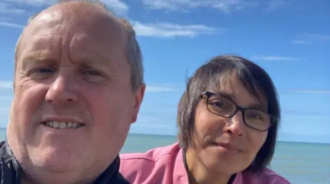 Andrew Powell Man and woman look at the camera, with a backdrop of the sea and blue skies. She has short dark hair and glasses and is wearing a pink blouse.