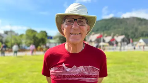 James Burstow is wearing a red t-shirt, with a bucket hat and wears glasses and is smiling in a field on a sunny day.