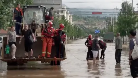 People stand on an excavator vehicle's front loader bucket in the middle of a flooded road.