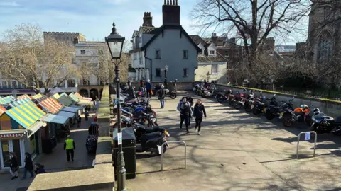A large concrete area holds around thirty motorbikes. To the left, the multi‑coloured roofs of Norwich Market stand out brightly, and in the distance you can see the cathedral and an old building.
