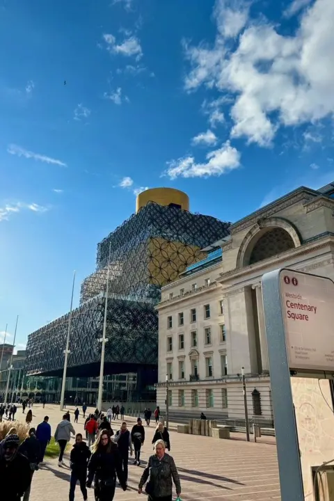 @BiteYourBrum A modern building made up of a stack of four rectangular staggered sections all clad with interlocking metal rings, stands next to a 1930s art deco building to the right of it. People are walking across a plaza in front and a sign reads "Centenary Square".