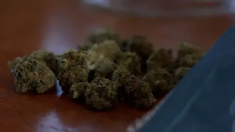 Dried cannabis flower is being poured out of a blue packet onto a wooden table.