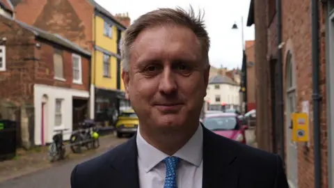 Shaun Whitmore/BBC Ed Garratt stands on a street with several red bricked buildings surrounding it including a yellow one. He has short light hair with some grey in it. He wears a navy suit, white shirt and a tie. 