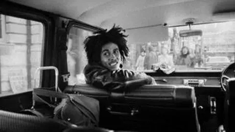 Dennis Morris A black-and-white photograph shows Bob Marley seated in a vehicle, turned partly toward the camera as he leans over the backrest of a seat, with people visible through the windows and the vehicle interior around him.