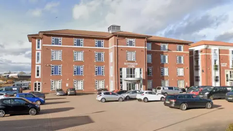Google Scotswood House in Thornaby. It is a large four-storey building made from red bricks. Similar buildings stand on either side of it and a large car park is located in front of the main entrance.