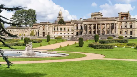 English Heritage Witley Court in Worcestershire, a stately home. It is in the Italianate building style and is set back from extensive gardens, with well-manicured lawns, trees and bushes. There is a fountain in the foreground.
