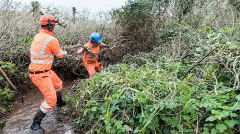 Somerset Rivers Authority Workmen wearing orange high vis clothing and hard hats are shown clearing what appears to be a small stream. They are standing in running water and are moving a large branch. There is lots of green vegetation around the water. 