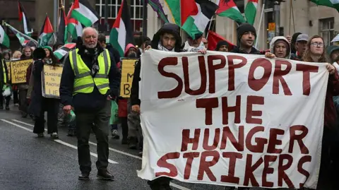 Dozens of protesters carrying Palestinian flags are seen in Liverpool. At the front, several people hold a large banner which has the words 'Support the hunger strikers' painted on it