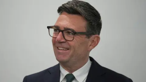 Andy Burnham looks off camera to the left. He is wearing a dark suit, white shirt and dark tie. 