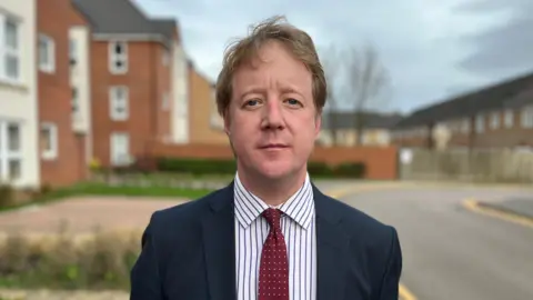 Paul Bristow faces the camera, wearing a dark suit, striped shirt and red spotty tie. He is stood on a pavement outside a block of flats. 