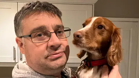 BBC A middle-aged man with grey hair and glasses holding a ginger and white dog next to his face. They are in front of some white kitchen cabinets