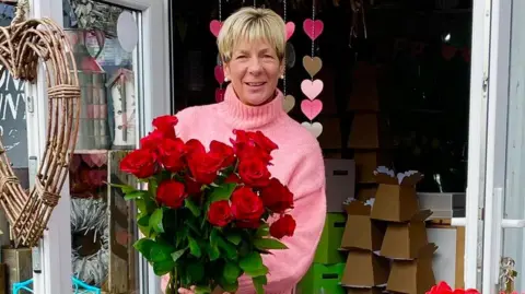 A woman with short blonde hair wearing a pink jumper and blue jeans. She is hold a big bouquet of red roses that have a green stem. 