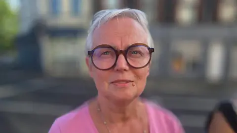Romana Nowak has short grey hair and is wearing a pink top and brown round frame glasses.