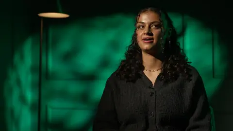 Ruby Greensides sits facing the camera in a studio interview, wearing a dark cardigan, with green light patterns behind her.