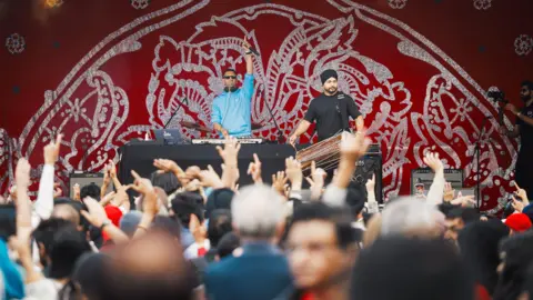 Glasgow Life A DJ and musician stand onstage in front of a cheering crowd - the DJ is wearing a blue top and has one hand raised in the air while at the decks