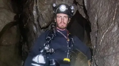 George Linnane inside a cave and looking at the camera. He is wearing a wetsuit and a safety helmet. 