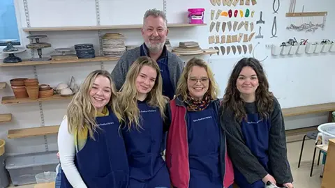 A family of a mother, father and their three adult daughters sit in a pottery studio, smiling at the camera. They all wear matching navy aprons over their clothes. 