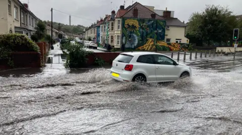 Anthony Ward/BBC A white car drives through floodwater close to the M32 in Eastville, Bristol. A row of terraced houses is visible, as is the Lockleaze Tower in the background