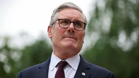 Starmer looking past the camera. He is wearing black framed glasses, a black suit jacket, white shirt and a burgundy tie. He has grey hair. The background is blurred but he appears to be outside with trees behind him.
