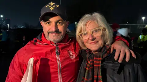 BBC A man and woman smile for the camera, but both look emotional. The man has his arm around the woman, who has blonde bobbed hair and cream coloured glasses. The man wears a baseball hat, has a greying beard, and wears Hull KR kit.