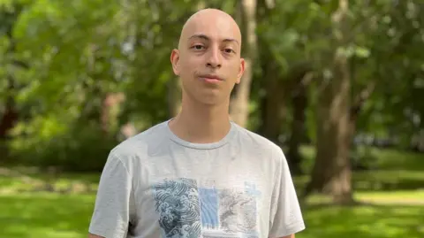 A bald man with black eyebrows looks at the camera with a slight smile. He's wearing a grey T-shirt with waves pictured on it 