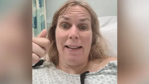 Joanne Graham A woman with shoulder length blonde hair and dressed in a hospital gown holds her thumb up to the camera