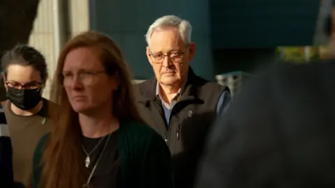 BBC Ian Wilkinson wearing a black vest and spectacles, with a head of white hair. He is walking, surrounded by several others.