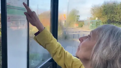 Jannette Ward looks up and points to the paper timetable at her bus stop in Herefordshire as the tracking app can be unreliable