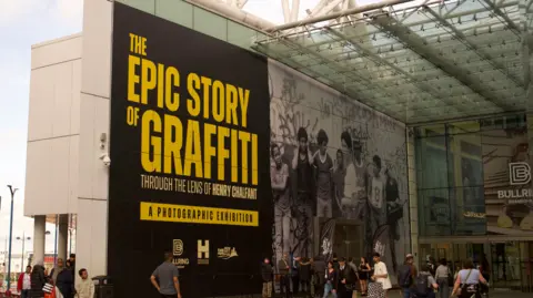 A giant image with the words 'The Epic Story of Graffiti: Through the lens of Henry Chalfant' in yellow on the side of an entrance to a modern shopping centre.