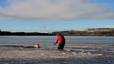 Frankie, with a swimming cap and goggles, swims through a channel in the icy Sweethope Lough. Coach Fenwick Ridely is standing in the water, knee deep, in a red coat and fluffy hat and monitoring her progress. 