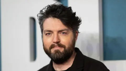 A man with dark hair and a dark wearing wearing a black shirt.