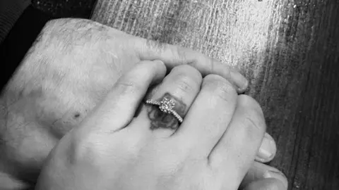 Charlotte Busby Two hands photographed. Charlotte lays her hand over Matthew's. She has a single-stone ring on her ring finger. Charlotte has a tattoo of a crown on her ring finger which is visible under the band. 