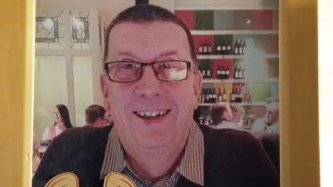 Rod Walsh is smiling at the camera. He is wearing glasses and a jumper over a shirt. He is in a restaurant and there are people in the background. The photo of Rod is in a frame.