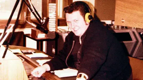Brian Shuttlewood wearing a dark jacket sat with headphones on in front of a microphone and mixing board.