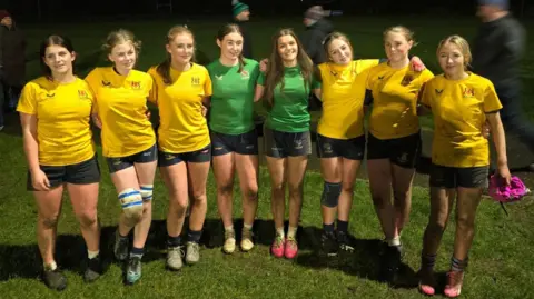 Neil Campbell A team of eight girls all standing in a line with arms around each other. They are wearing yellow jerseys and two wearing green jerseys in the middle. They are standing on the pitch after a game, covered in mud. 