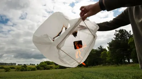 A person is holding a white paper sky lantern in a field with trees in the backdrop. Visible orange flames are rising.