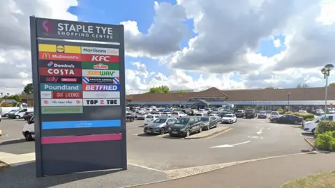 Google The car park at Staple Tye Shopping Centre in Harlow. On the left is a grey sign which reads "Staple Tye Shopping Centre", and the sign lists the shops and restaurants at the centre