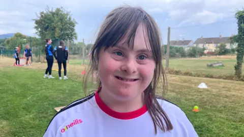 Kate has long brown hair with a fringe that falls to her eyebrows. She is smiling and wearing a white top with a pink collar and black stripes across the shoulders.