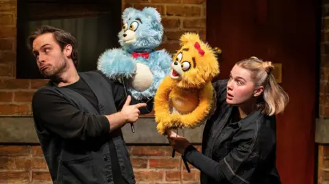 Two performers manipulate blue and yellow furry puppet characters during a scene from Avenue Q, with a brick wall set visible behind them.