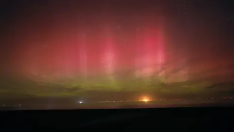 BBC Weather Watchers/andyaurorahunter A night-time landscape of the North Sea. There are wind turbines far away and the Northern Lights have made the sky turn pink and yellow, and there are several stars.