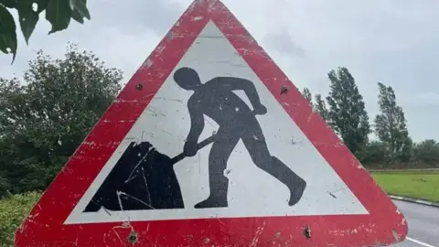 A red triangular roadworks street sign with a silhouetted man shown digging. A road with trees and grassland are behind the sign.