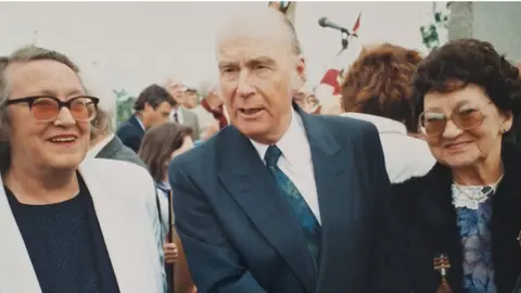 Liam Duggan Shows two women wearing glasses standing either side of President Patrick Hillery, who has a with receding hairline and is wearing a blue, double-breasted suit, white shirt and blue and green floral tie.