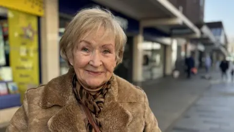 Paula smiles as she poses in front of a row of high street shops. She has short fair hair and wears an animal print scarf around her neck. She also wears a sandy-coloured coat.