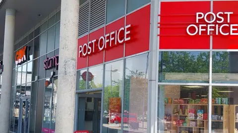 Post Office The outside of a post office, with red and white signage, glass windows and items inside the store. A door is open to the left. 