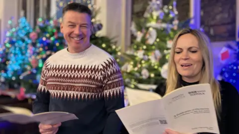 Jon Wright/BBC Luke Deal and Sarah Lilley singing from service-sheets inside a church with Christmas trees in the background.