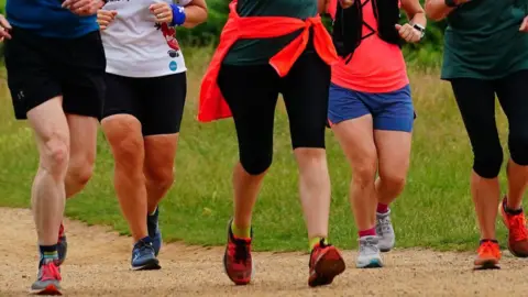 Runners head along a path in a park. Only their lower halves can be seen. They are all wearing shorts and running trainers.